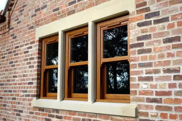 A set of three oak effect uPVC vertical sliding windows with double-glazed panels, framed by a red-brick wall and light stone surrounds.